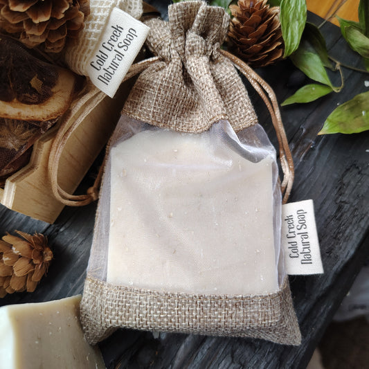 A bar of natural soap wrapped in a burlap sack with a transparent window, displaying the soap inside. The sack is tied with a string and has a label that reads 'Cold Creek Natural Farm'.