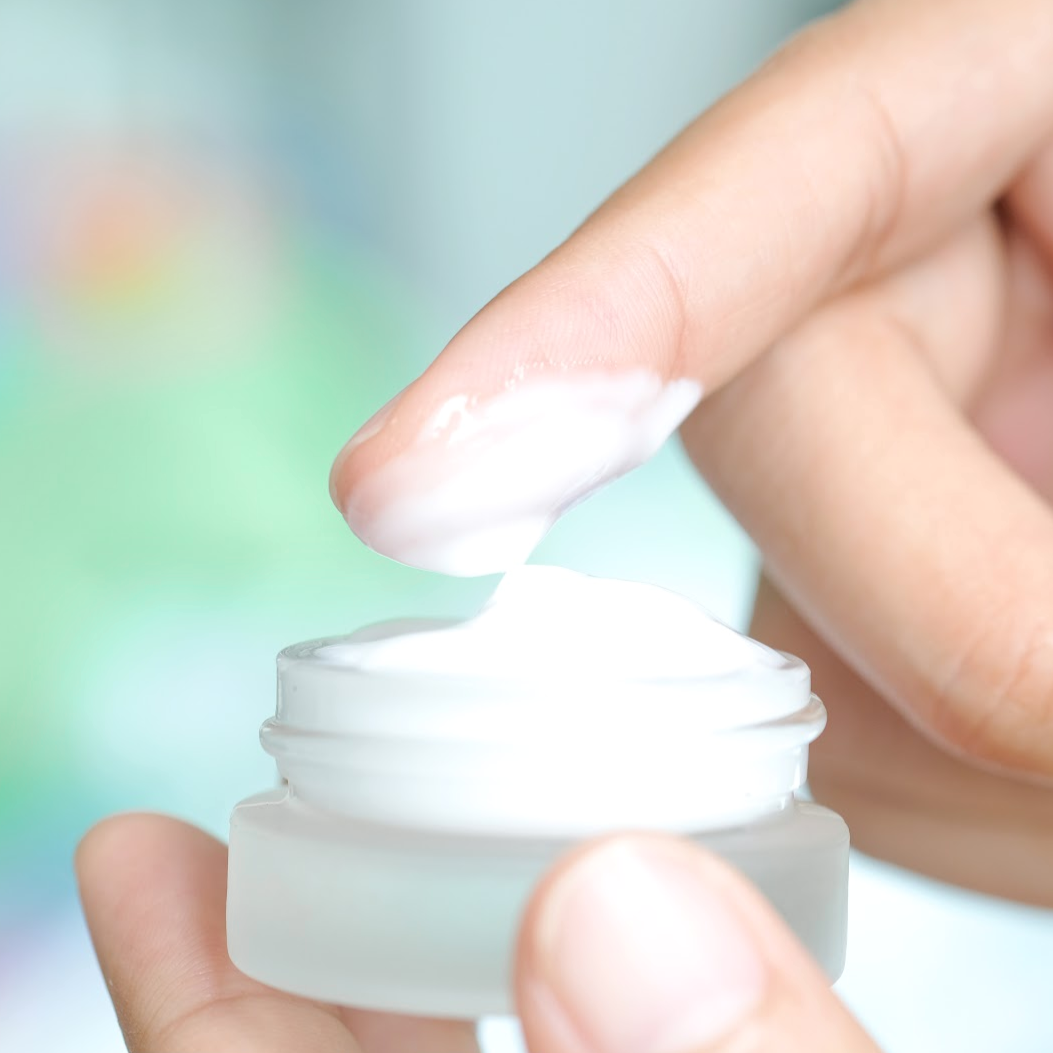 Hand applying cream from a small white container with a blurred colorful background