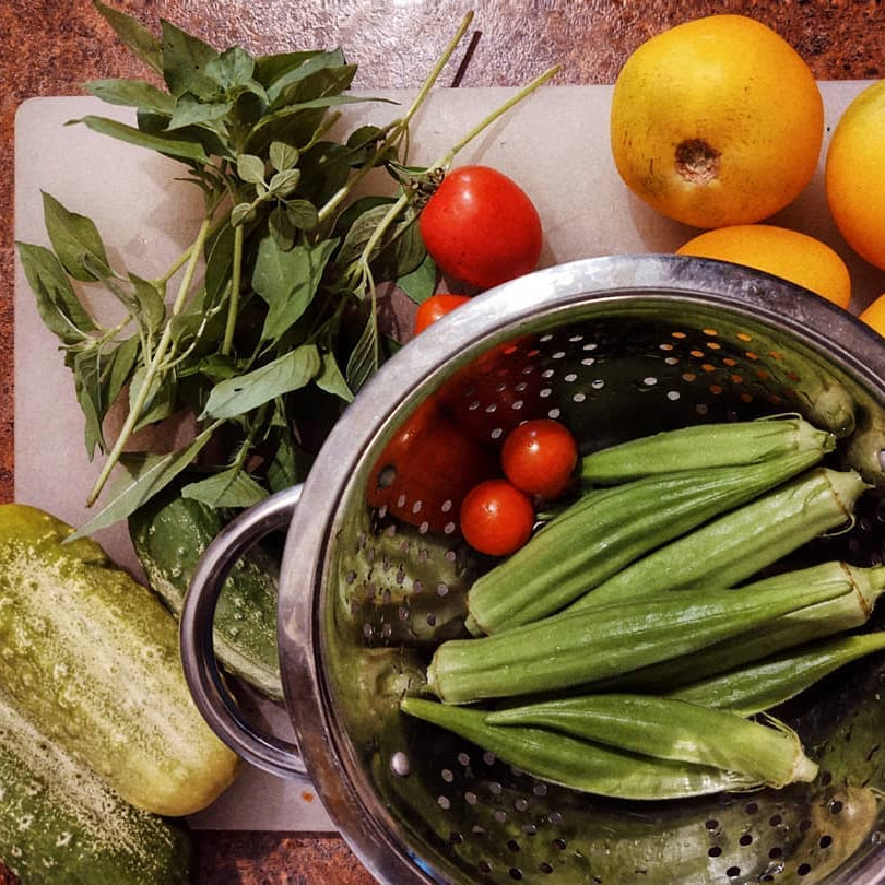 Colander with okra, tomatoes, and herbs on a cutting board with oranges.